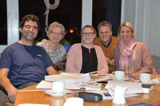 Fünf Personen, drei Frauen und zwei Männer, sitzen an einem Tisch mit Dokumenten, Büchern und Kaffeetassen. Sie lächeln in die Kamera. Der Raum ist gemütlich beleuchtet, im Hintergrund sind Fenster mit Dekoration und Pflanzen.